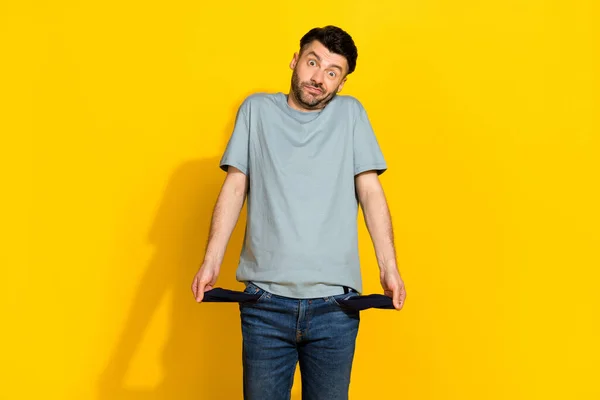 Photo of young funny excited stressed man frustrated confused problems showing his empty pockets poverty no money isolated on yellow color background.