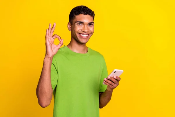 年轻英俊学生的照片 成功的自由撰稿人博主拿着手机 在黄色的背景下 Okey 的标志 微笑着漂亮的广告 空旷的空间 与世隔绝 — 图库照片