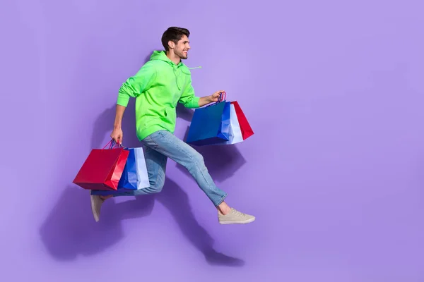全长照片 穿着绿色便宜货的时髦兴奋男子跳过空旷空间孤立的紫色背景 — 图库照片