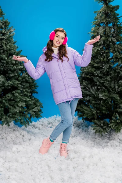 明るい魅力的な女の子の縦の肖像は アウターウェアを身に着けている耳暖かい青の色の背景に隔離された雪を立つ — ストック写真