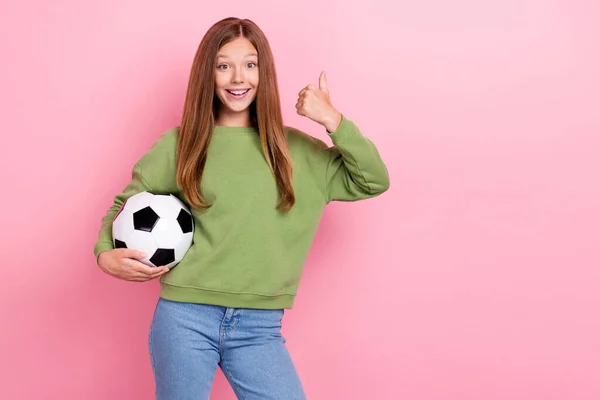 照片上 穿着绿色运动衫的有自信的 闪亮的女生拿着足球大拇指在空旷的空间里撑起了孤立的粉色背景 — 图库照片