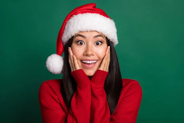 Closeup photo of young korean woman wear red pullover nice santa claus hat touch cheeks shock black friday sale isolated on green color background.