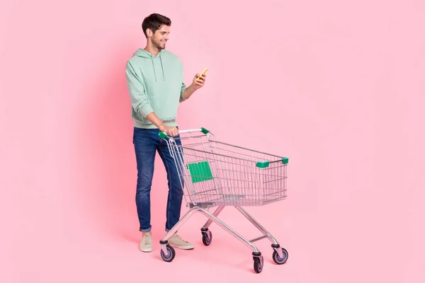 正面时尚男人拿着购物车的全长照片阅读表购买产品超市空旷的空间隔离在粉红色的背景下 — 图库照片