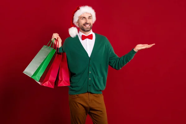 笑顔の写真サンタクラスヘルパー男は緑のカーディガンホールドパイルショッピングモールバッグヤシの木は 赤の色の背景に隔離された新しいオファーを実証 — ストック写真