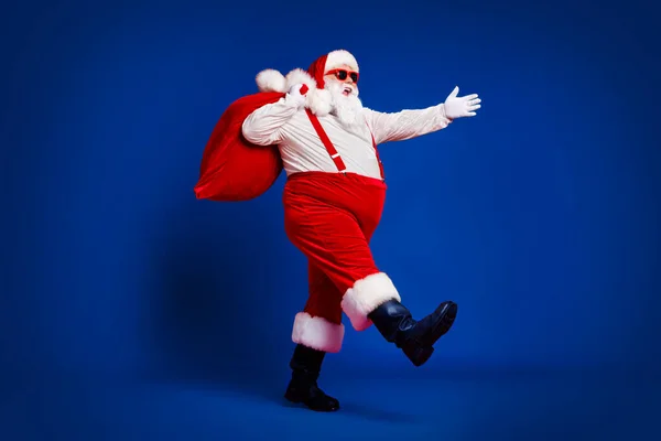 全身上下尽收眼底 他那迷人的欢快欢快滑稽的圣诞老人背着大袋大减价走向孤立明亮鲜亮的红色底色 — 图库照片