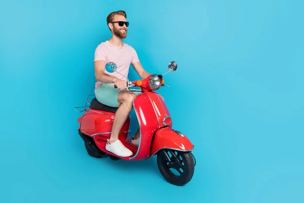 Full length photo of nice young man driving scooter summer sunglass tourist wear trendy pink clothes isolated on cyan color background.