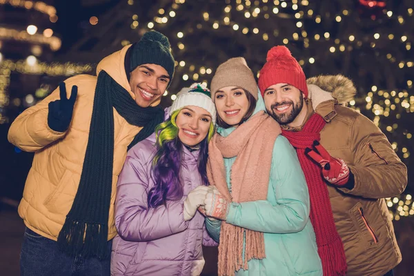 Portrait of positive friendly people hold hands demonstrate v-sign have fun evening tree lights garland outside.