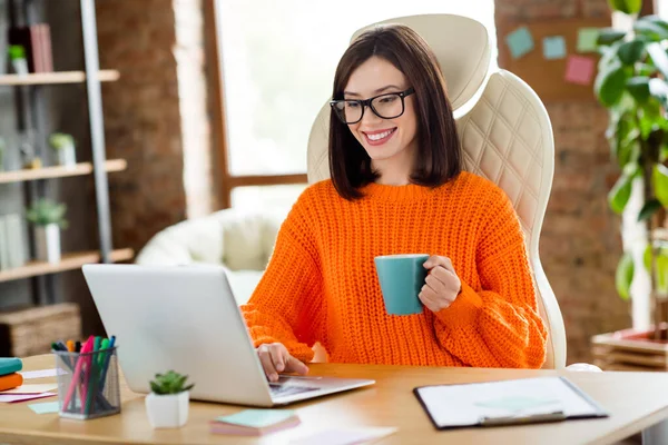 照片上快乐迷人的女孩她坐在皮椅上手握茶杯用上网本在室内写电子邮件工作站 — 图库照片
