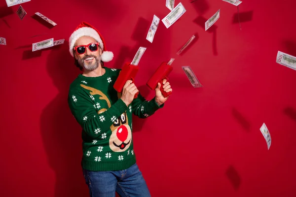 Portrait of positive crazy person hold money gun shoot dollar hundred banknotes bills isolated on red color background.