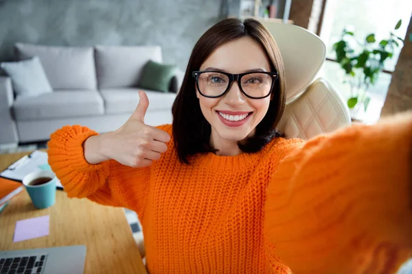 梦幻般自信的女士穿着橙色毛衣自拍的照片 在室内车间工作站展示大拇指 — 图库照片