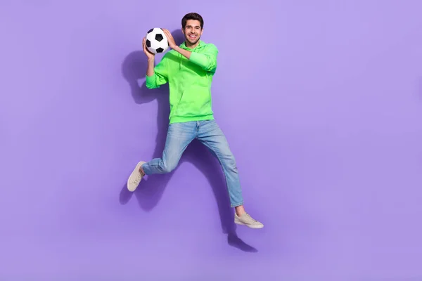 Full length photo of cheerful funky guy dressed neon sweatshirt jumping high playing football isolated violet color background.