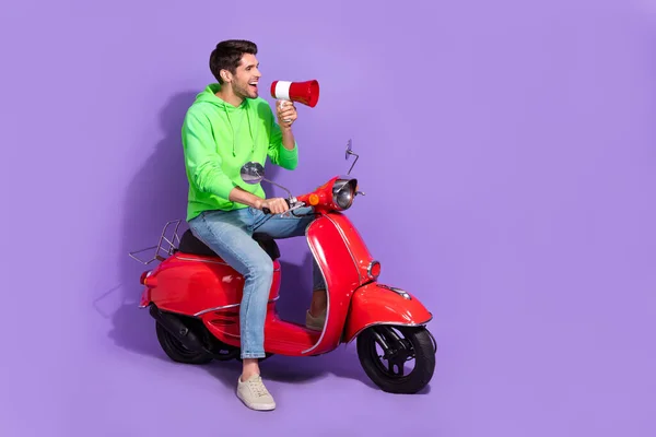 Full size photo of satisfied activist guy green hoodie hold loudspeaker look empty space on motorcycle isolated on violet color background.
