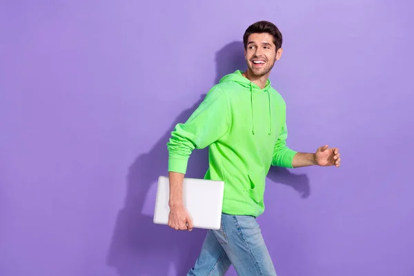 stock image Photo of positive attractive guy with brunet hairdo dressed green hoodie hold laptop look empty space isolated on purple color background.