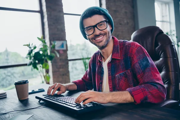 图为快乐微笑的编码员戴着帽子眼镜打字程序现代小玩意室内工作站阁楼的照片 — 图库照片