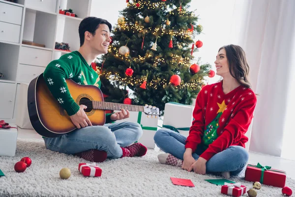 照片上的年轻人穿着华丽华丽的装饰毛衣 令人毛骨悚然的圣诞气氛弹吉他放松歌唱小夜曲清凉的地毯在室内冷杉树旁 — 图库照片