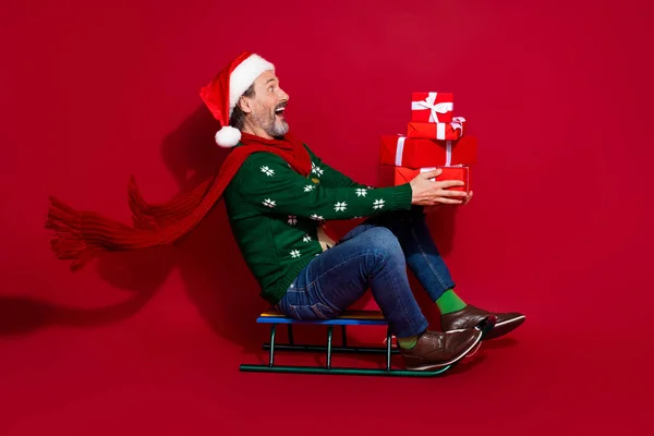 Full body profile portrait of astonished man sit ride sledge hold pile stack giftbox look empty space isolated on red color background.