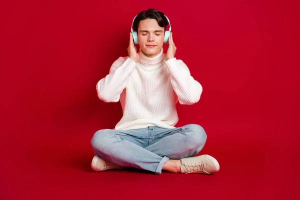 Full body photo of peaceful positive man sit floor closed eyes hands touch headphones isolated on red color background.