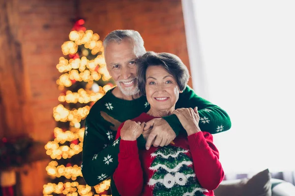 Closeup photo of two old seniors wear funny cozy ugly sweaters man hug his wife smiling wellbeing near christmas tree lights decor stay home.