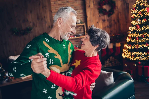 Side profile photo of old age couple seniors wear festive ugly sweaters hands together dancing winter mood music rhythm celebrate new year indoors.