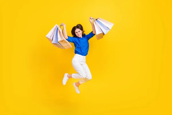 Full length photo of sweet lucky girl dressed blue shirt jumping high rising shoppers isolated yellow color background.