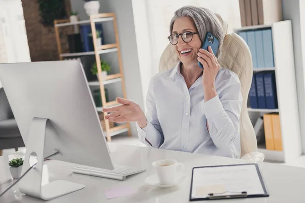 快乐的老年女律师的画像坐着说电话与同事在室内现代办公室交流 — 图库照片