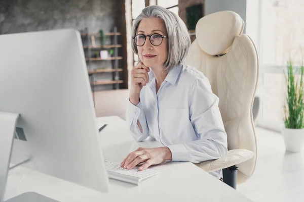 退休老妇人穿着白衬衫坐在明亮的现代工作站桌子上打字的照片 — 图库照片