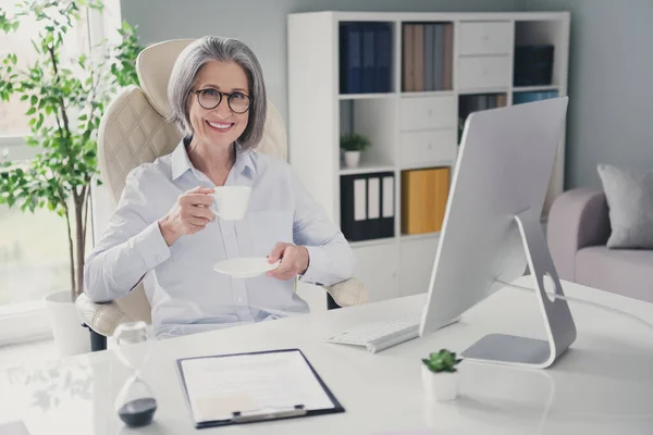 美丽而快乐的老年律师的画像坐着的女士把新鲜热咖啡杯和现代工作场所一起放在室内 — 图库照片