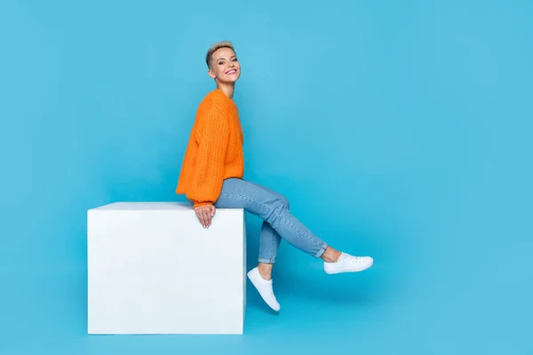 Full length photo of charming lady wear orange sweatshirt sitting platform having rest empty space isolated blue color background.