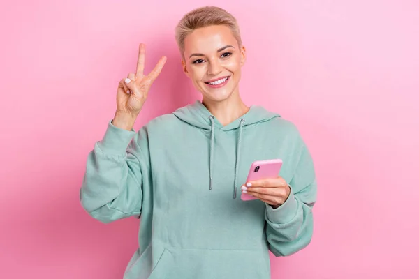 Photo of young cheerful friendly person instagram blogger on event showing v-sign peace hold phone stylish hoodie isolated on pink color background.