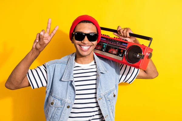Portrait of positive friendly person toothy smile hold boombox show v-sign isolated on yellow color background.