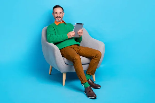成熟した素敵な男の完全な長さの写真座っているアームチェア読書Ebookタブレット着用トレンディーな緑の服に隔離された青の色の背景 — ストック写真