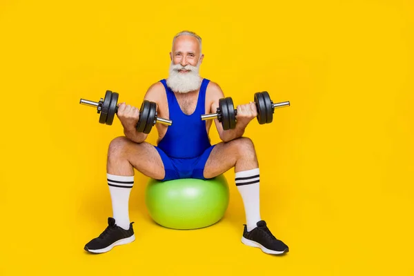 Full body length photo of old aged senior sitting gymnastics ball practicing dumbbells up powerful athlete isolated on yellow color background.