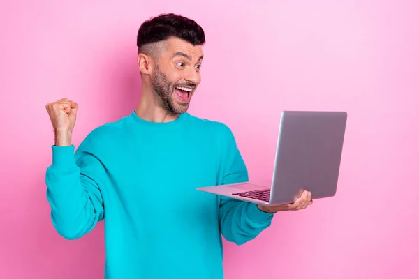 若い喜びに満ちた魅力的な男の写真青のトレーナーを着用してください拳アップホールドネットブックお祝いオンラインベット勝利ピンク色の背景に孤立 — ストック写真