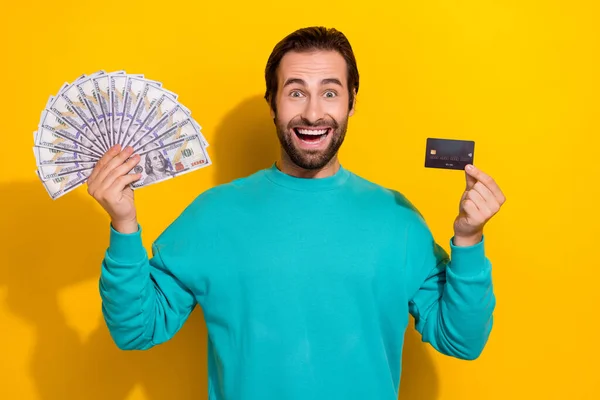 若い興奮した肯定的な笑顔のブルネットの髪の男の写真黒いプラスチックカードを保持彼の給料スタックドル黄色の背景に隔離 — ストック写真