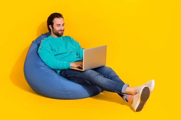 Full length photo of cheerful cool guy dressed teal outfit sitting bean bag typing gadget empty space isolated yellow color background.