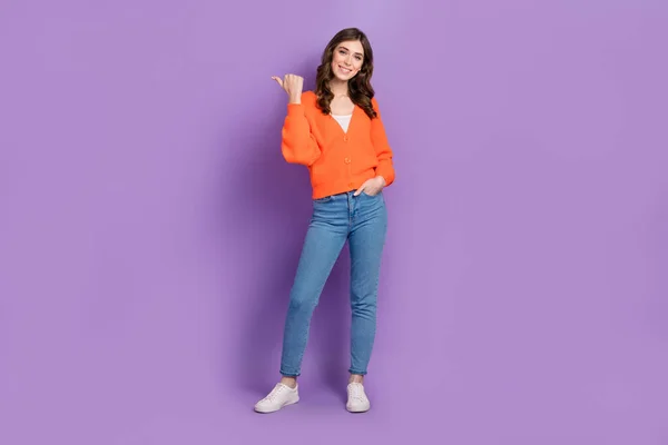 オレンジ色のカーディガンスタイリッシュなジーンズを身に着けている陽気な女性の完全な長さの写真の幹部直接親指モックアップ紫の色の背景に孤立便利なヒント — ストック写真