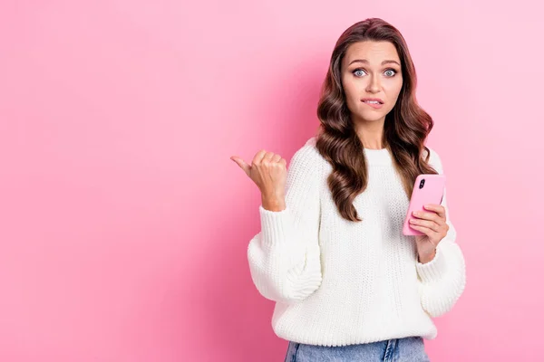 Photo of pretty nervous person biting lips hold telephone direct finger empty space isolated on pink color background.