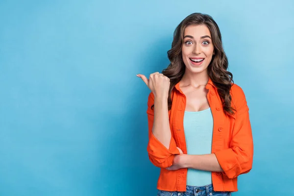Photo of confident excited lady wear orange jacket smiling pointing thumb empty space isolated blue color background.