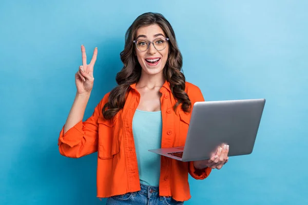 Photo of sweet excited lady wear orange jacket eyeglasses showing v-sign typing modern device isolated blue color background.