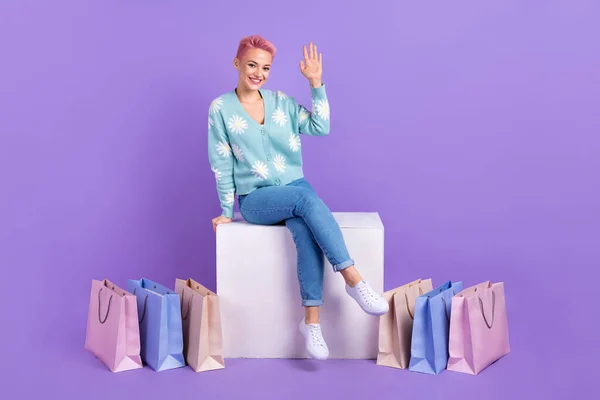 Full body length photo of young satisfied lady sit white box platform wave palm hello symbol near much shop bags isolated on purple color background.