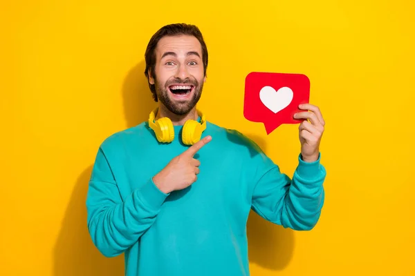 Photo of excited pretty man wear turquoise clothes headphones pointing love heart card isolated yellow color background.