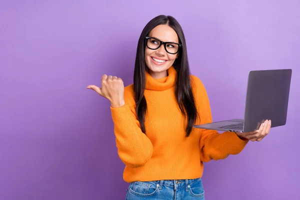 Stock image Photo of young business office worker woman wear orange jumper direct finger look empty space hold laptop offer isolated on purple color background.