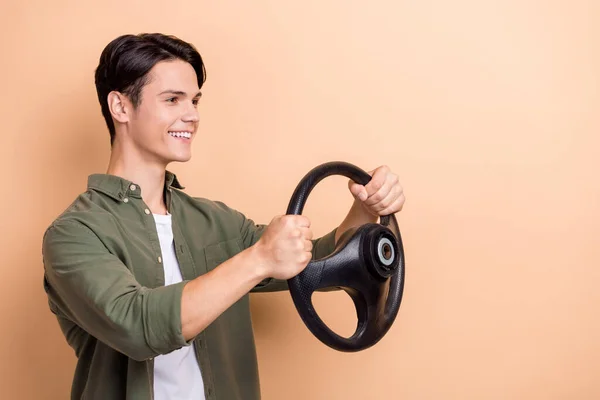 Photo of optimistic cool guy with brunet hair dressed khaki shirt hold steering wheel look empty space isolated on beige color background.