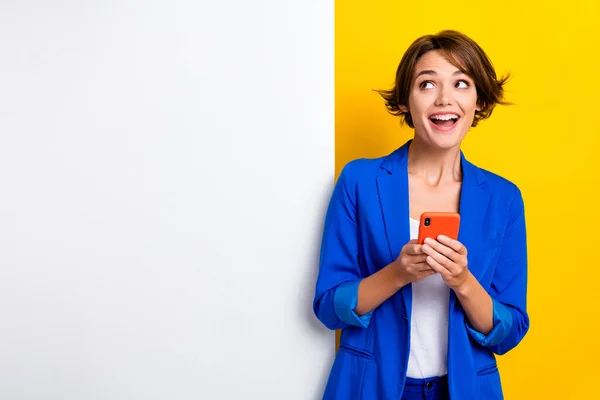 Photo of funny excited lady dressed blue blazer typing device looking white wall placard empty space isolated yellow color background.