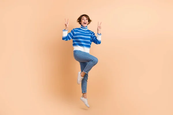 Full length photo jumping lady friendly blogger show fingers v-sign victory symbol peace greetings blue turtleneck isolated on beige color background.