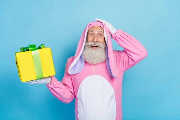 Portrait of speechless impressed man dressed pink rabbit costume hold present gift box arm on head isolated on blue color background.