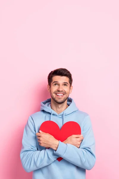 Vertical photo of dreamy sweet man wear blue hoodie looking up empty space embracing hugging heart isolated pink color background.
