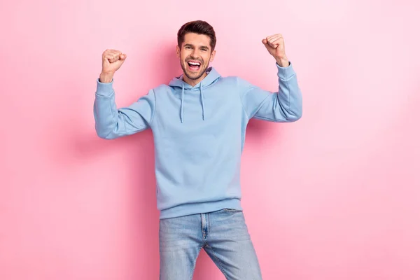 Photo of pretty lucky man wear blue hoodie screaming rising fists smiling isolated pink color background.