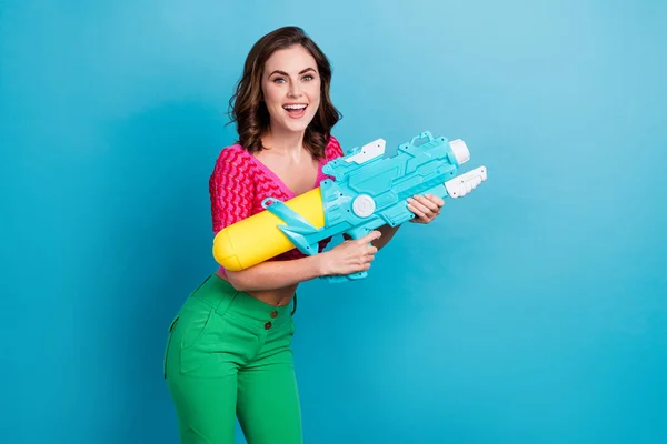Photo of optimistic good mood gorgeous girl with curly hairdo dressed pink top holding water gun isolated on blue color background.