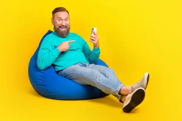 Full body photo of attractive grandpa sit beanbag excited point gadget dressed stylish cyan garment isolated on yellow color background.